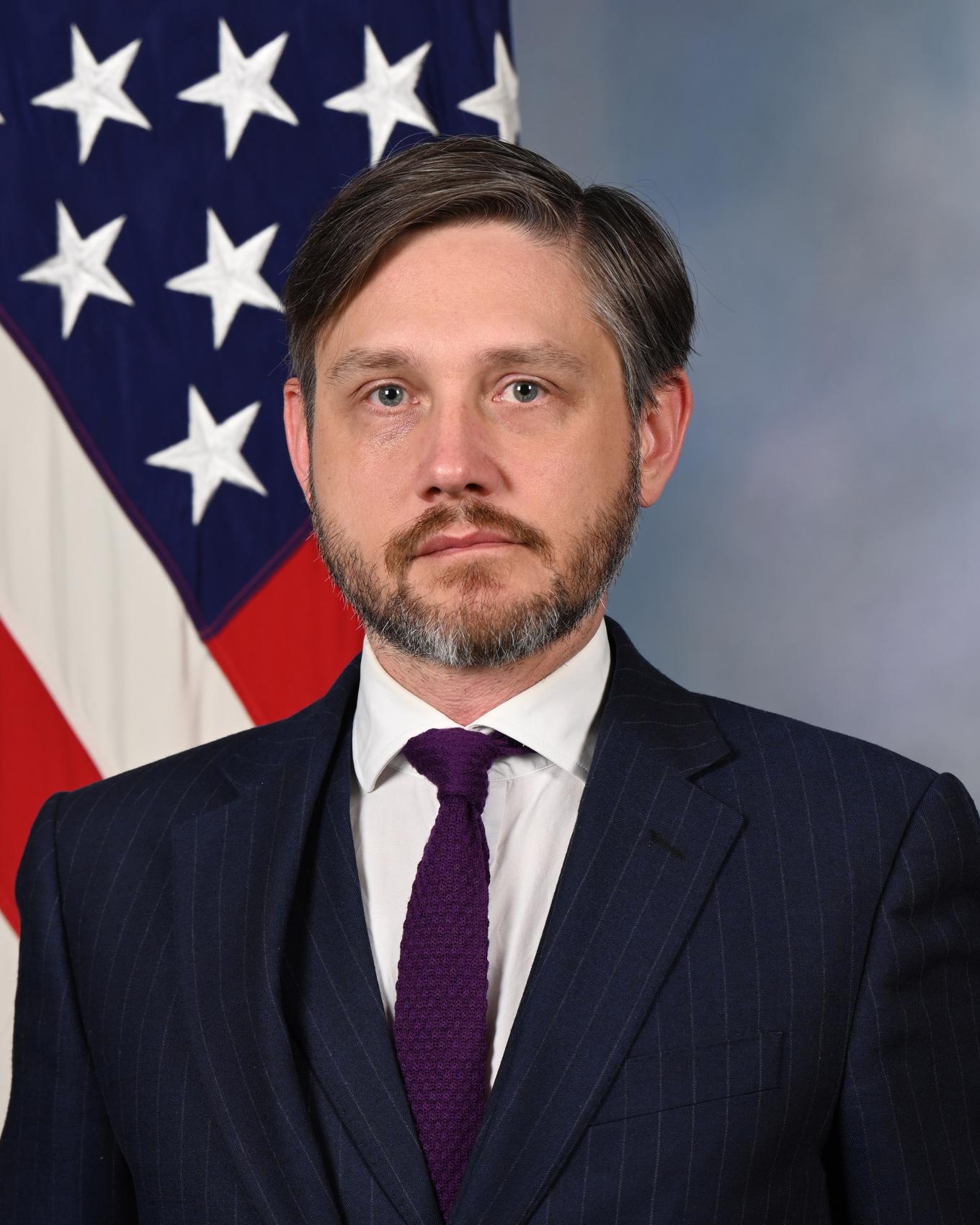 Dark haired man in navy blue suit in front of the American flag