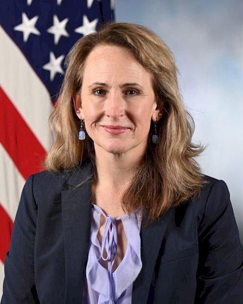 light brown haired woman in grey blazer in front of the American flag